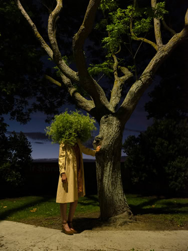 Photo by Florence Charvin Photography of A person wearing a tan coat stands at night beside a tree, their head obscured by a large, leafy green plant.