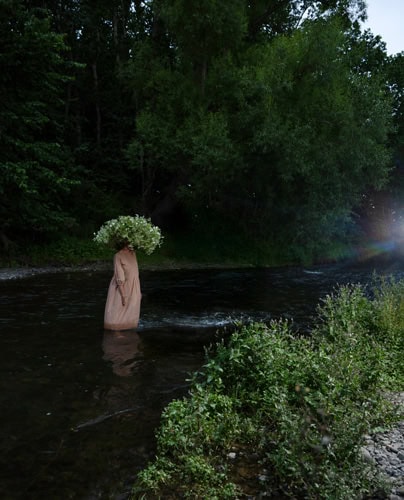 Photo by Florence Charvin Photography of A person in a long brown garment stands in a shallow river, holding or wearing a large bouquet of flowers that obscures their head; dense trees are in the background.