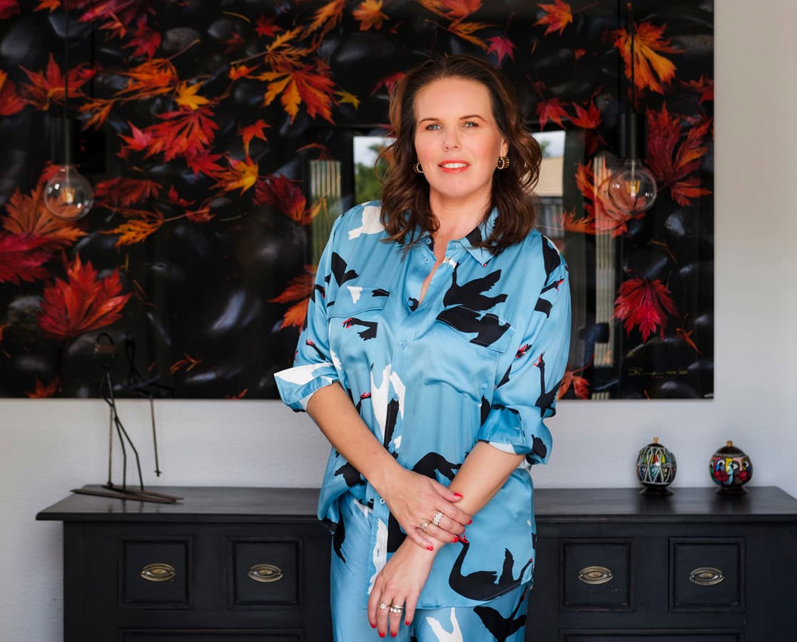 Photo by Florence Charvin Photography of A woman in a blue patterned outfit stands in front of a black chest of drawers and a wall art piece featuring autumn leaves.