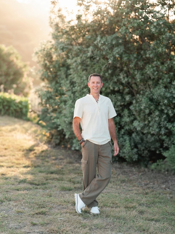 Photo by Florence Charvin Photography of A man in a light shirt and khaki trousers stands outdoors on grass near bushes, smiling at the camera in soft sunlight.