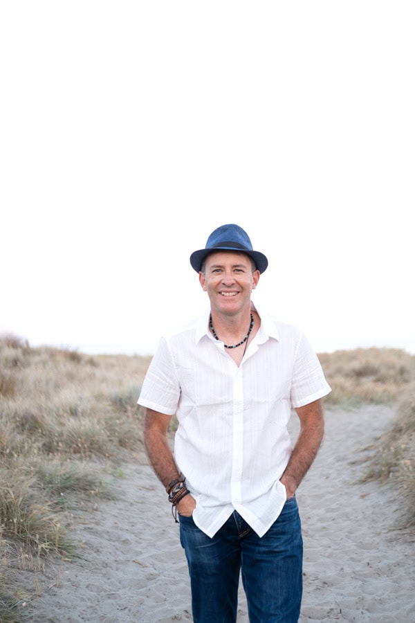 Photo by Florence Charvin Photography of A man wearing a white shirt, blue jeans, and a blue hat stands on a sandy path in a grassy dune area, smiling at the camera.