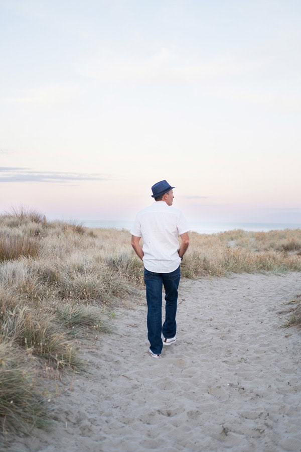 Photo by Florence Charvin Photography of A man wearing a blue hat, white shirt, and jeans walks alone on a sandy path through tall grass under a pastel sky.