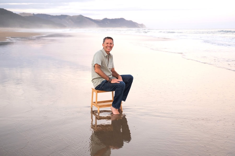Photo by Florence Charvin Photography of A man sits barefoot on a wooden chair on wet sand at the beach, with waves and distant mountains in the background.