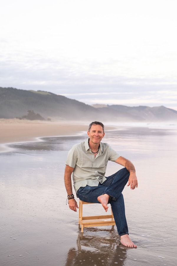 Photo by Florence Charvin Photography of A man sits barefoot on a wooden chair on a wet sandy beach, with misty hills and the sea in the background.