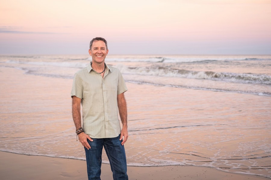 Photo by Florence Charvin Photography of A man stands on a beach at sunset, smiling at the camera. The sky is pastel-coloured and gentle waves approach the shore in the background.