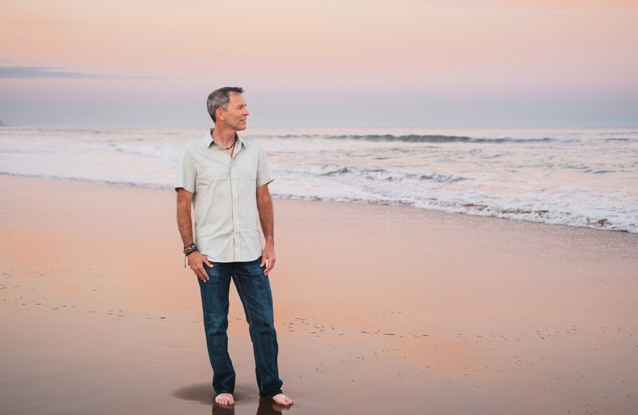 Photo by Florence Charvin Photography of A man stands barefoot on a beach at sunset, looking to his left. Gentle waves and a pastel-coloured sky are visible in the background.
