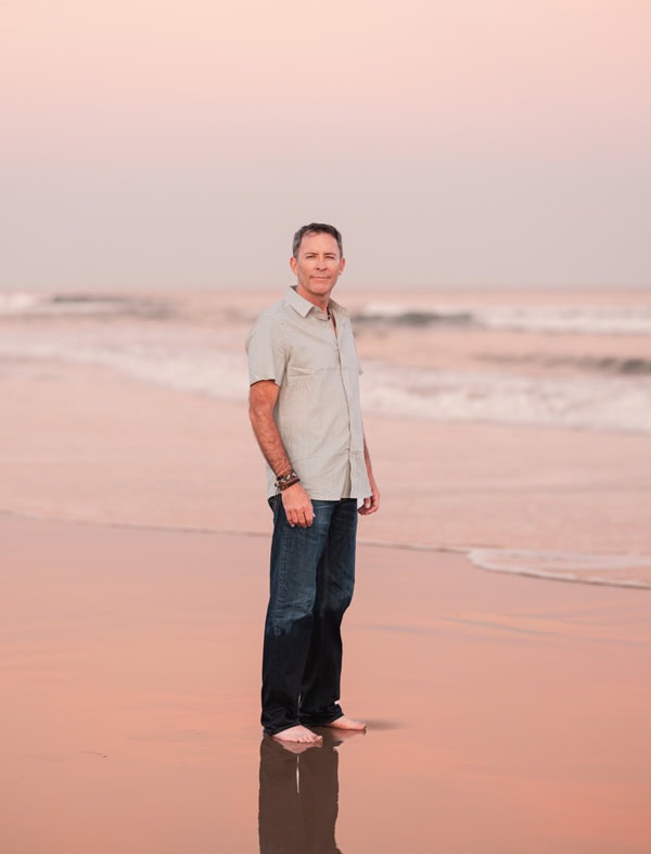 Photo by Florence Charvin Photography of A man stands barefoot on a sandy beach near the shore at sunset, with gentle waves and a pastel-coloured sky in the background.