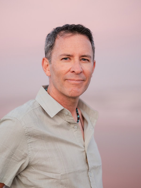 Photo by Florence Charvin Photography of A middle-aged man with short grey hair, wearing a light-coloured shirt, stands outdoors against a soft pink background, looking at the camera.
