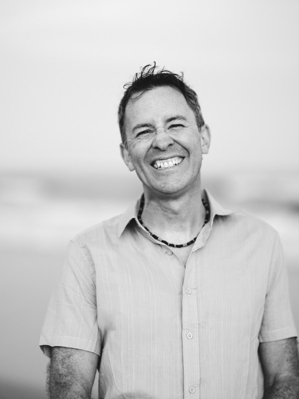 Photo by Florence Charvin Photography of A man wearing a short-sleeve button-up shirt and necklace smiles broadly outdoors with a blurred background. The image is in black and white.