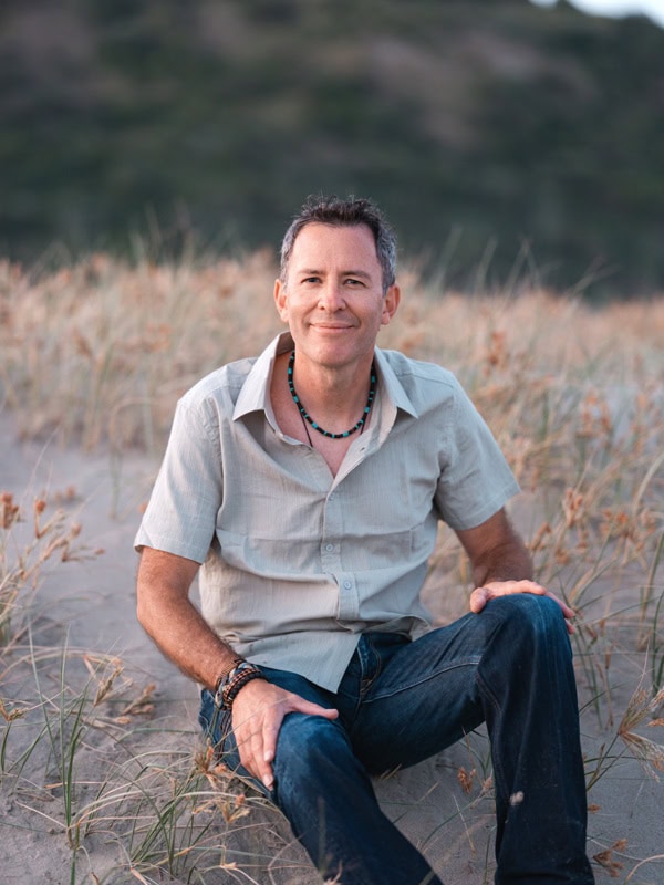 Photo by Florence Charvin Photography of A man with short hair and a beaded necklace sits on sandy ground among dry grass, wearing a short-sleeved shirt and jeans, with blurred greenery in the background.