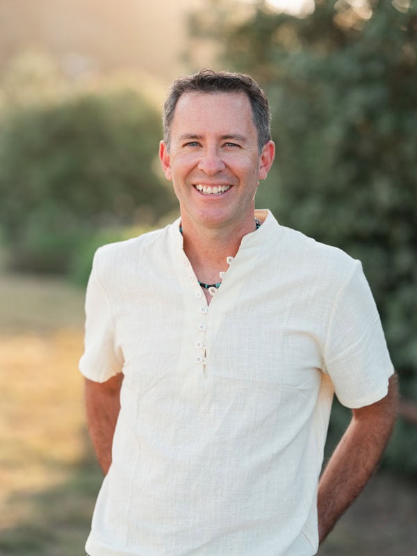 Photo by Florence Charvin Photography of A man wearing a short-sleeved, light-coloured shirt stands outdoors, smiling at the camera with trees and sunlight in the background.
