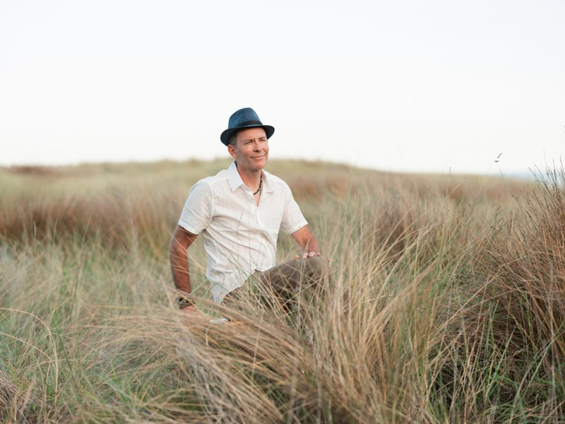 Photo by Florence Charvin Photography of A man wearing a short-sleeved shirt and a black trilby hat sits in tall grass, with a blurred field and sky in the background.