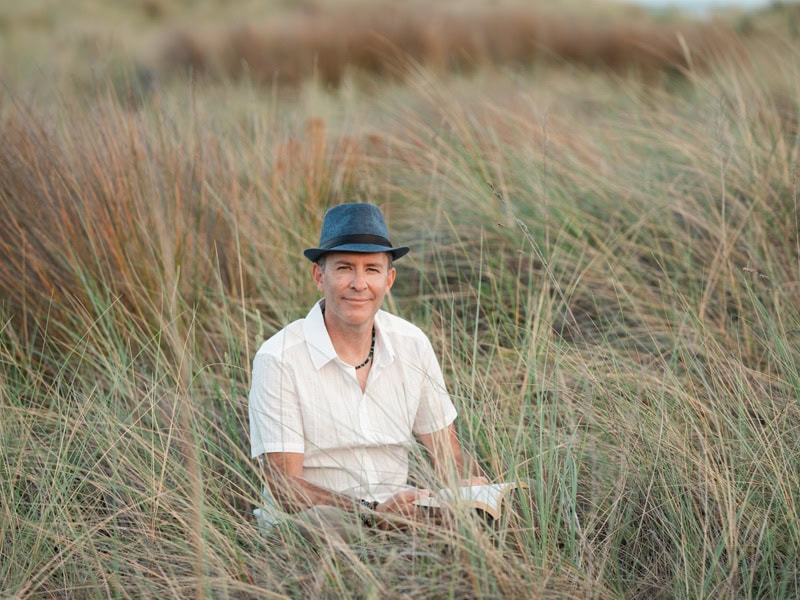 Photo by Florence Charvin Photography of A man wearing a blue hat and white shirt sits in tall grass, holding an open book and smiling at the camera.