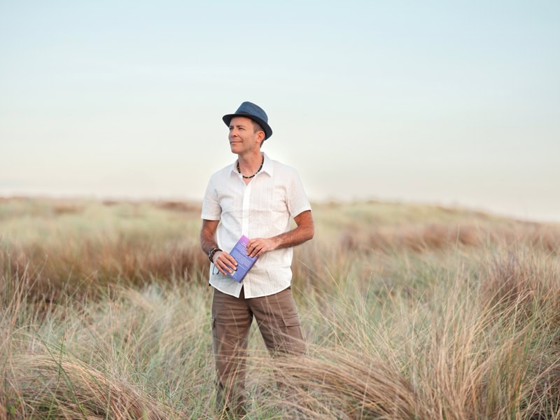 Photo by Florence Charvin Photography of A man wearing a short-sleeve shirt and hat stands in tall grass, holding a book, with a blurred landscape in the background.