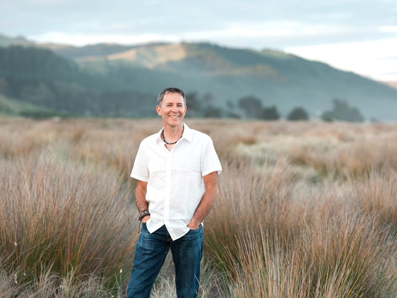 Photo by Florence Charvin Photography of A man stands outdoors in a grassy field with mountains in the background, wearing a light-coloured short-sleeve shirt and jeans, smiling with his hands in his pockets.