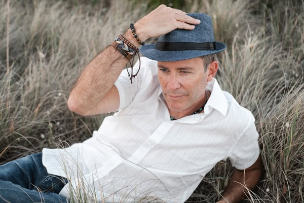 Photo by Florence Charvin Photography of A man in a white shirt and blue hat reclines in tall grass, holding his hat with one hand and wearing several bracelets on his wrist.