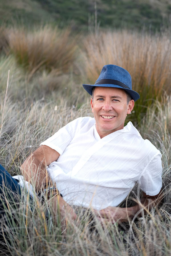 Photo by Florence Charvin Photography of A man wearing a blue hat and white shirt lies smiling in tall grass outdoors with blurred plants in the background.