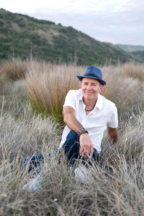 Photo by Florence Charvin Photography of A man wearing a blue hat and white shirt sits in tall grass with hills and cloudy sky in the background.