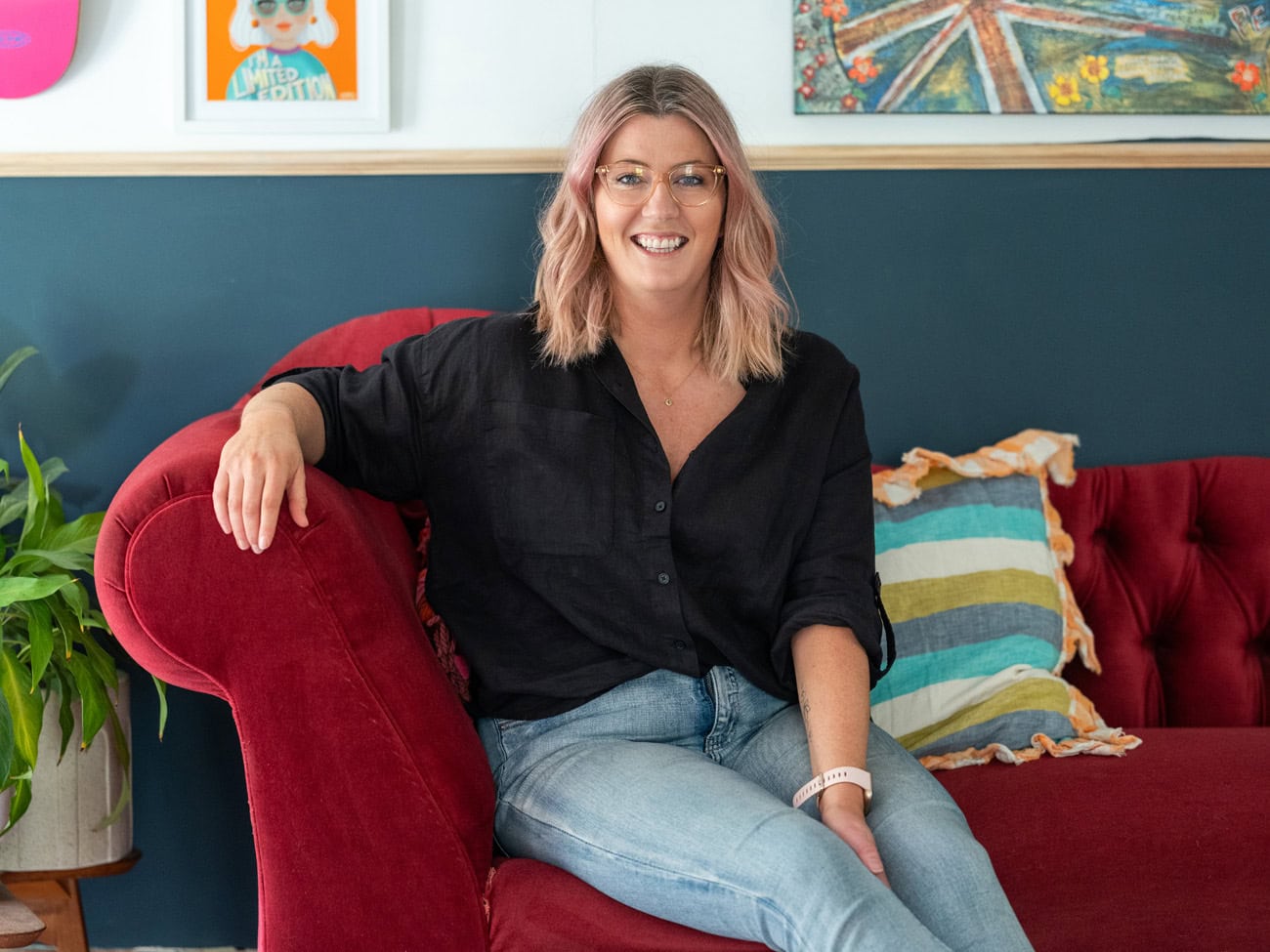 Photo by Florence Charvin Photography of A woman with shoulder-length pink hair and glasses sits on a red sofa, smiling, with her arm resting on the backrest. Colourful artwork and a striped cushion are visible in the background.