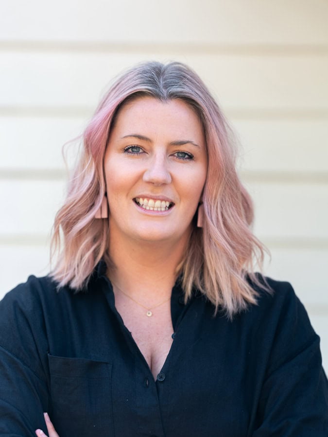 Photo by Florence Charvin Photography of Woman with shoulder-length light pink hair wearing a black top, standing in front of a light-coloured wall and smiling at the camera.