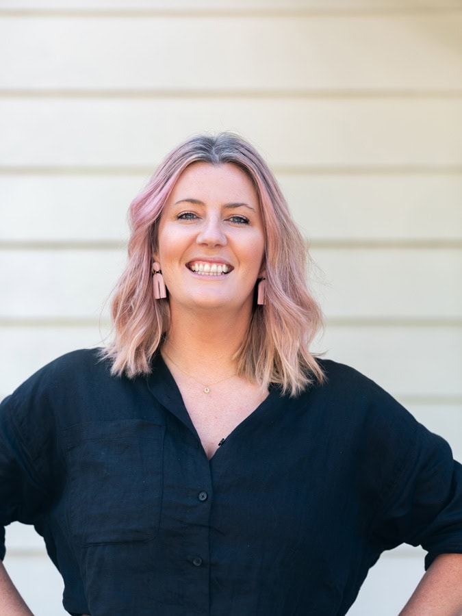 Photo by Florence Charvin Photography of Woman with shoulder-length pink hair wearing a black shirt and pink earrings, smiling and standing in front of a light-coloured horizontal panel wall.