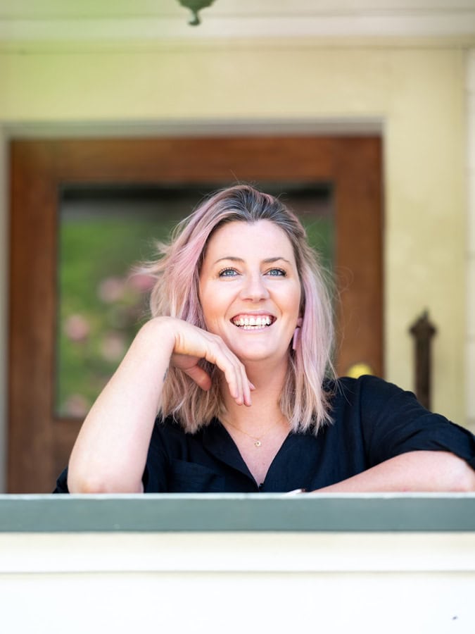 Photo by Florence Charvin Photography of A woman with shoulder-length fair hair and a black top smiles while sitting on a veranda, resting her chin on her hand.