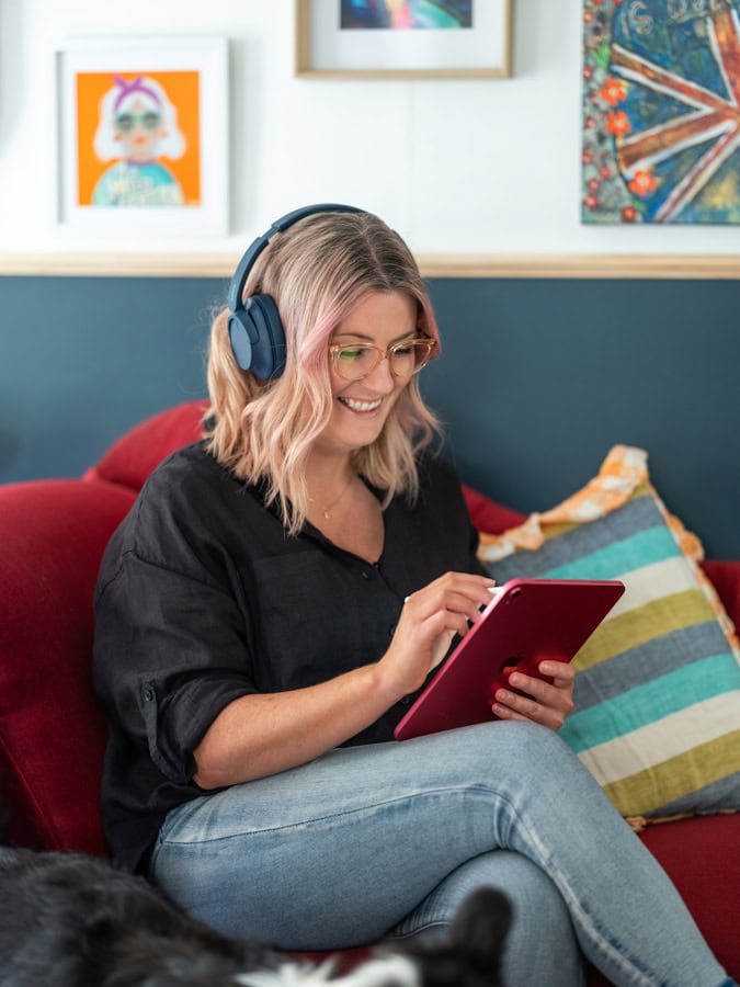 Photo by Florence Charvin Photography of A woman wearing headphones and glasses sits on a red sofa, smiling whilst using a tablet. Colourful artwork and a striped cushion are visible in the background.