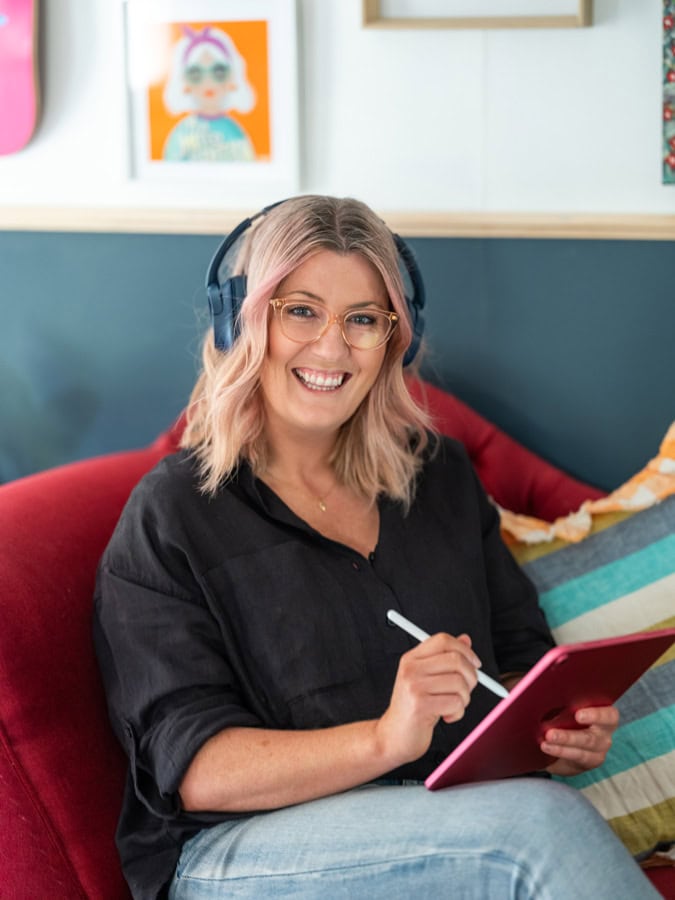 Photo by Florence Charvin Photography of A woman with shoulder-length blonde hair and glasses sits on a red chair, wearing headphones and using a stylus on a pink tablet, smiling at the camera.
