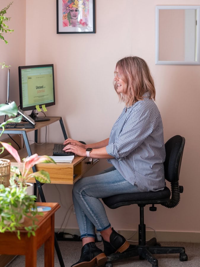 Photo by Florence Charvin Photography of A woman sits at a desk working on a computer in a home office, surrounded by plants and wall decor.