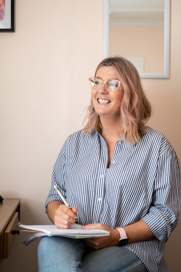 Photo by Florence Charvin Photography of A woman with shoulder-length hair and glasses smiles while writing in a notebook, seated indoors in a casual setting.