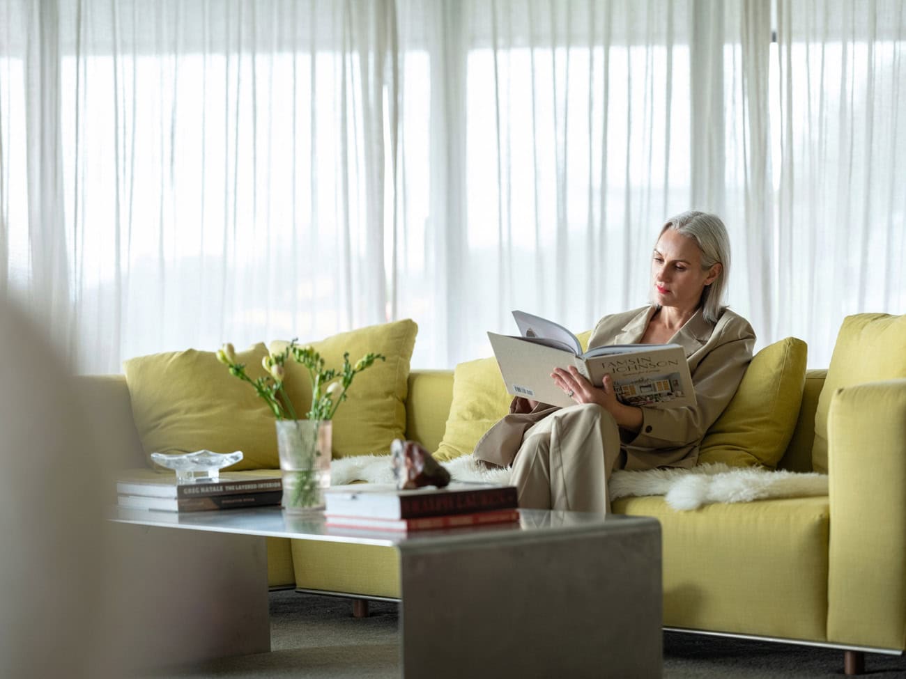 Photo by Florence Charvin Photography of A woman sits on a yellow sofa reading a large book, with her feet up on the cushion; a table with books and a vase of flowers is in front of her.