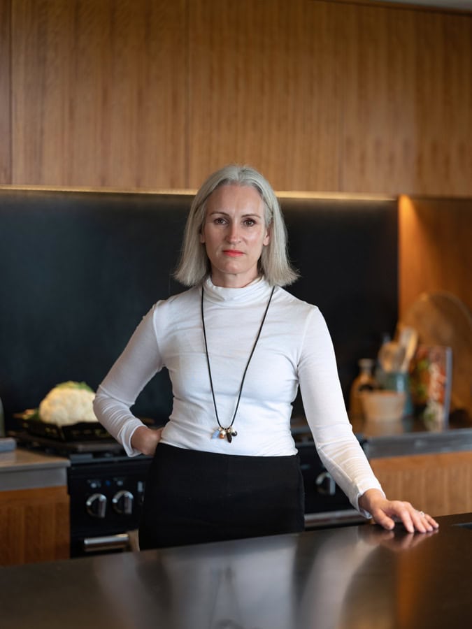 Photo by Florence Charvin Photography of A woman with grey hair stands in a modern kitchen, wearing a white polo neck and black skirt, with one hand on the worktop and the other on her hip.