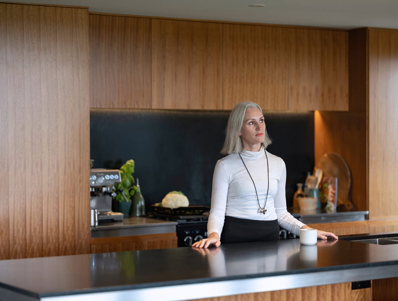 Photo by Florence Charvin Photography of A woman with grey hair wearing a white polo neck stands at a kitchen counter with a mug, surrounded by modern wooden cupboards.