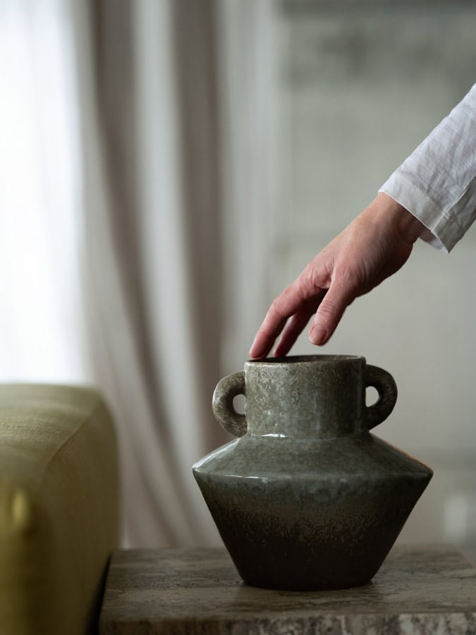 Photo by Florence Charvin Photography of A hand reaching towards a ceramic vase with two handles, placed on a stone table next to a green sofa in a softly lit room.