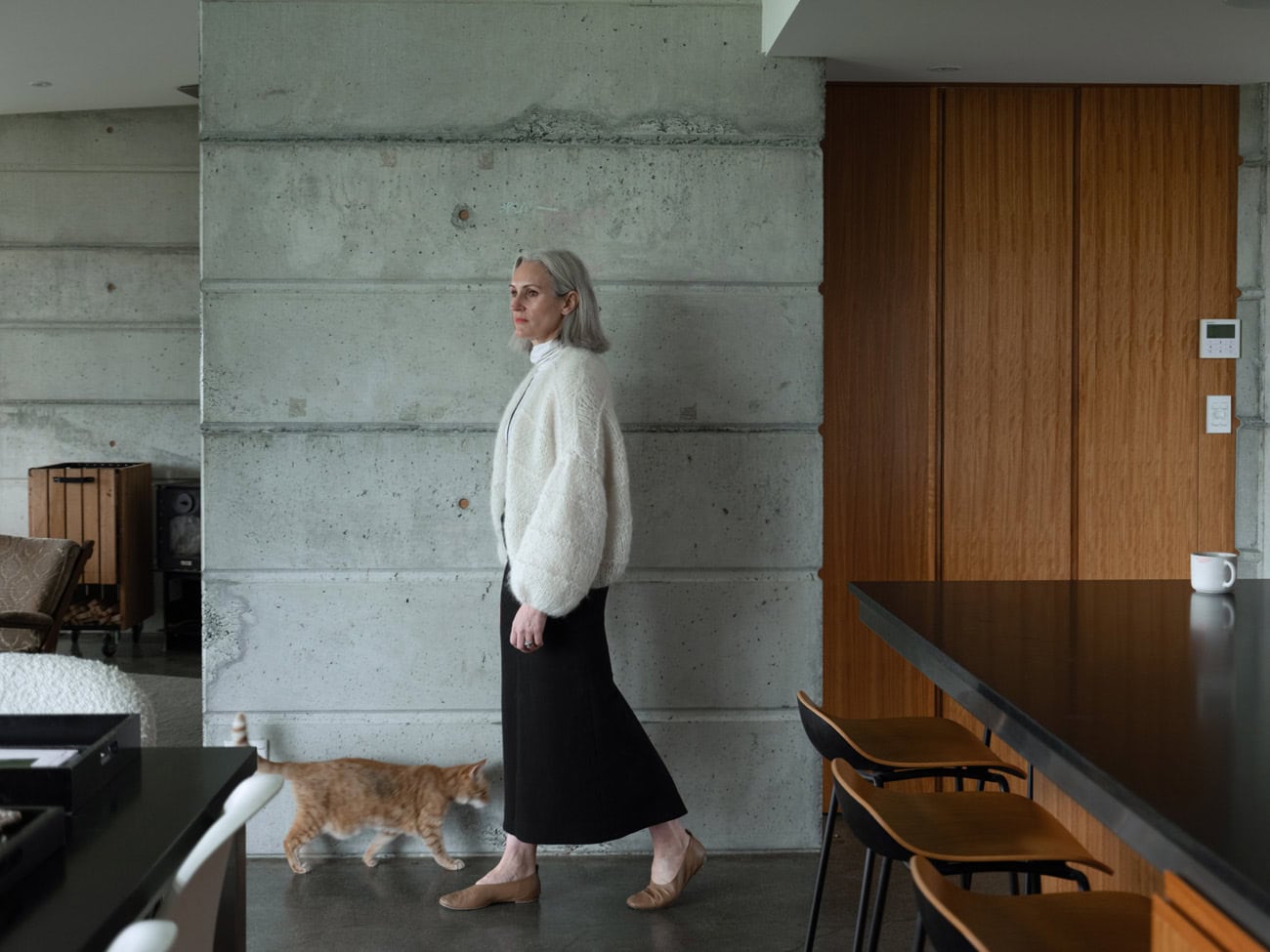 Photo by Florence Charvin Photography of An older woman with grey hair stands indoors near a concrete wall, wearing a white jumper and black skirt. A small orange cat walks by her feet. Modern furniture is visible.
