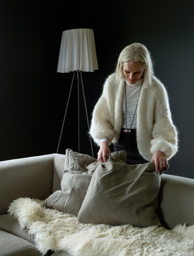 Photo by Florence Charvin Photography of A person with long blonde hair arranges cushions on a beige sofa in a room with dark walls and a floor lamp in the background.