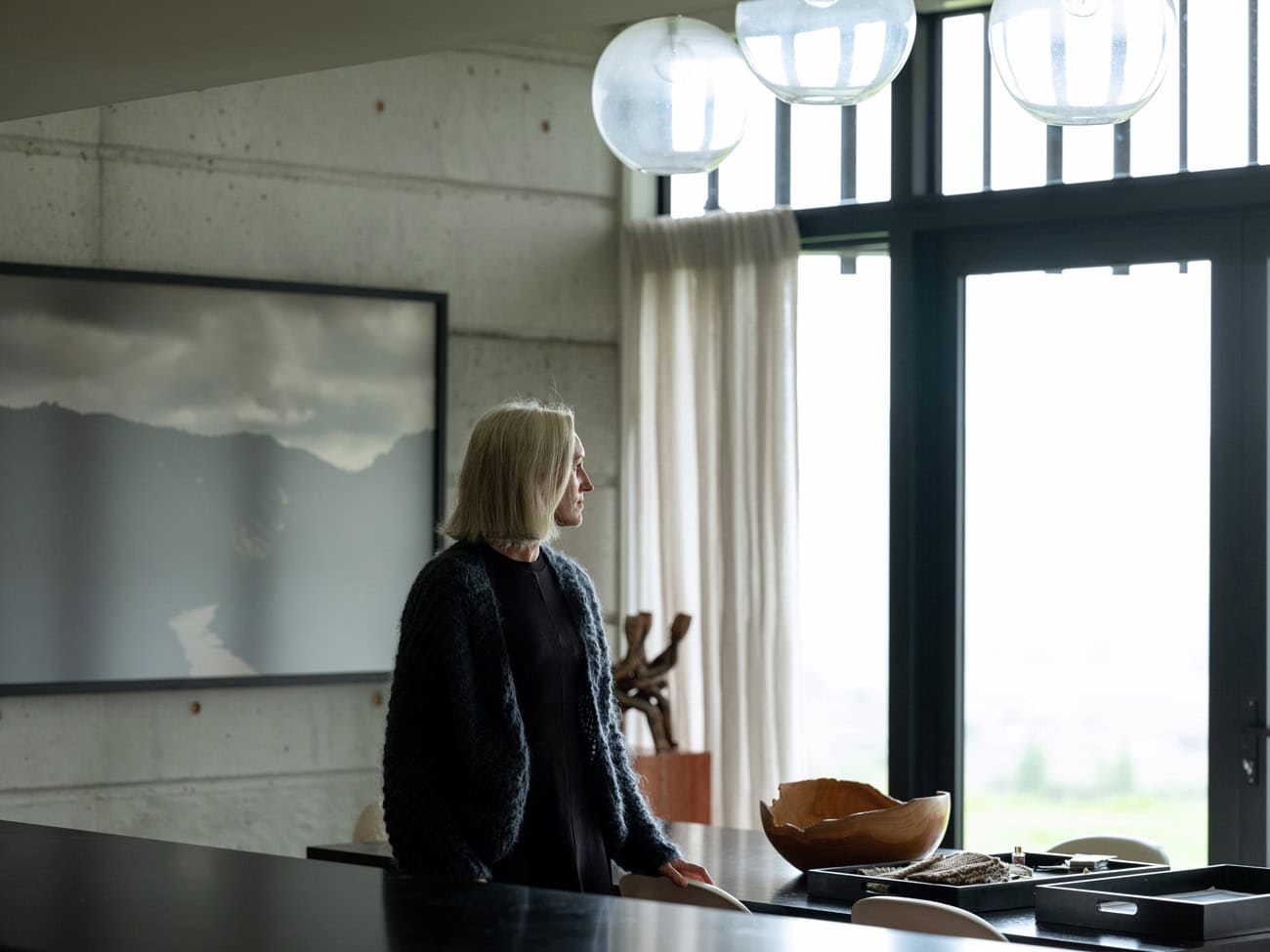 Photo by Florence Charvin Photography of A woman with grey hair stands in a modern kitchen, looking out a large window with sheer curtains. A wooden bowl and decorative items are on the counter.