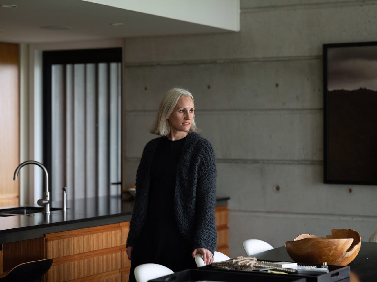 Photo by Florence Charvin Photography of A woman with grey hair stands in a modern kitchen with concrete walls, wooden cupboards, and a black worktop, looking to the side.