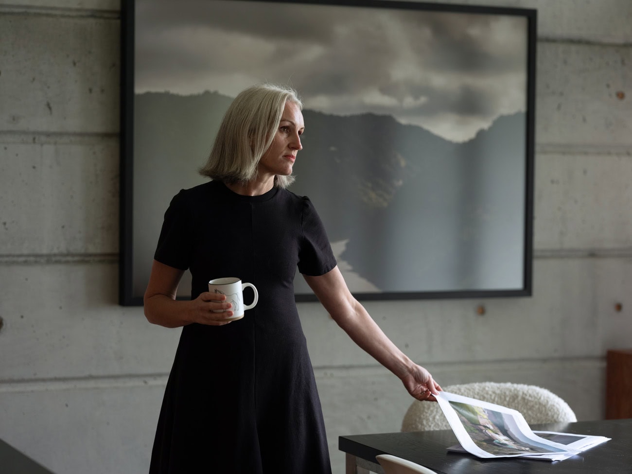 Photo by Florence Charvin Photography of A woman with grey hair in a black dress holds a mug and looks aside while reaching for a magazine on a table, with a landscape photo framed on the wall behind her.