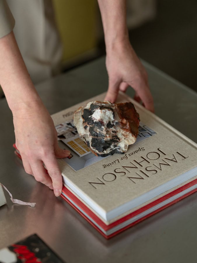 Photo by Florence Charvin Photography of A person places a large rock on top of a hardback book titled "Tamsin Johnson Spaces for Living" on a metal surface.