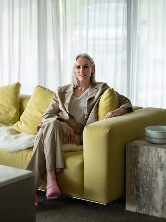 Photo by Florence Charvin Photography of A woman with fair hair wearing a beige suit and pink slippers sits on a yellow sofa in a living room with sheer white curtains.