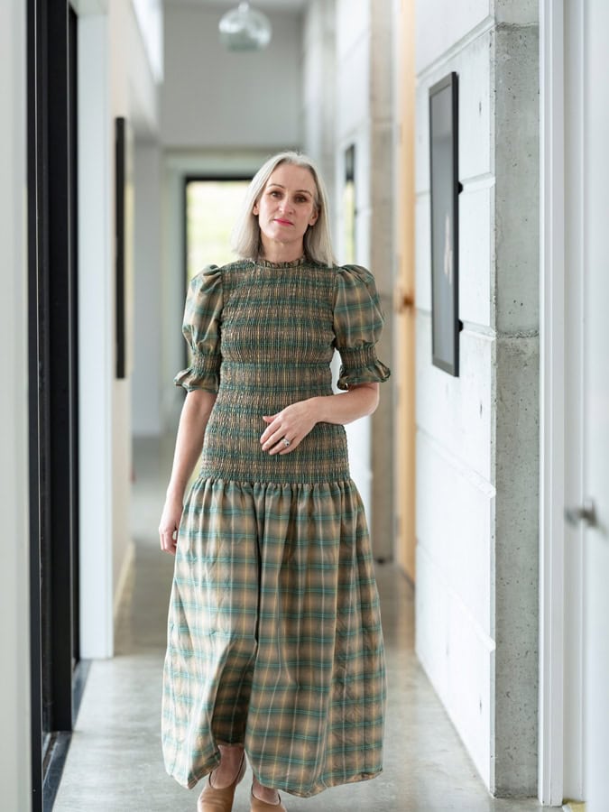 Photo by Florence Charvin Photography of A woman in a green checked dress with puffed sleeves stands in a modern corridor with concrete walls and framed pictures.