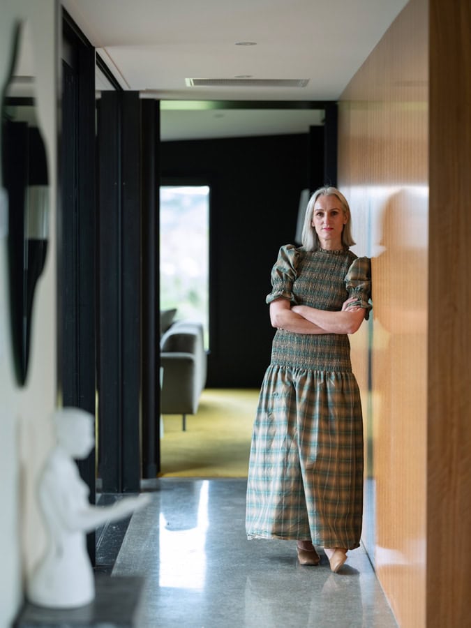 Photo by Florence Charvin Photography of A woman in a checked dress stands with arms crossed in a modern corridor, with a grey statue visible in the foreground.