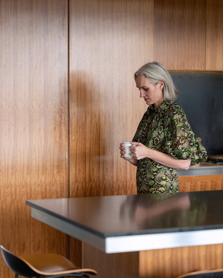 Photo by Florence Charvin Photography of A woman with grey hair stands in a kitchen, holding a white mug. She wears a green patterned dress and is next to a wooden wall and a black worktop.