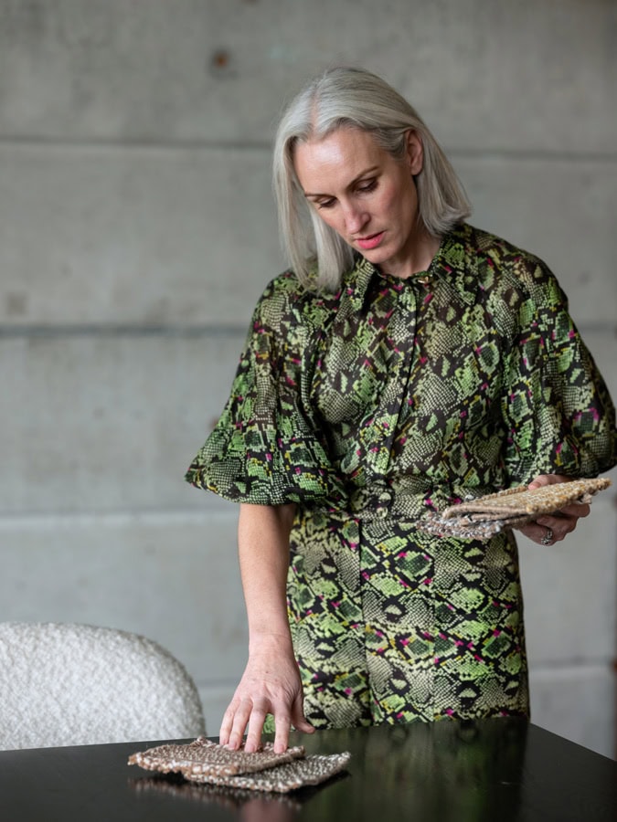 Photo by Florence Charvin Photography of A woman with grey hair wearing a green snakeskin-patterned outfit stands by a table, examining two textured fabric samples.