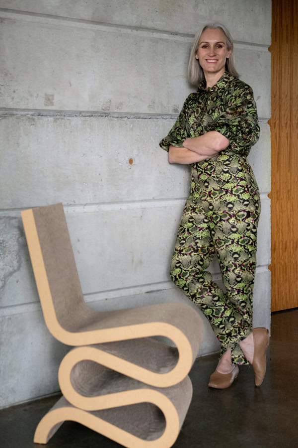 Photo by Florence Charvin Photography of A person with grey hair, wearing a green patterned playsuit and tan shoes, stands against a concrete wall beside a modern, curvy chair.