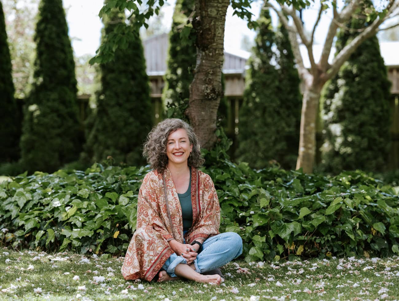 Photo by Florence Charvin Photography of A woman with curly grey hair sits cross-legged on grass, smiling, wearing a patterned shawl and jeans, with greenery and trees in the background.