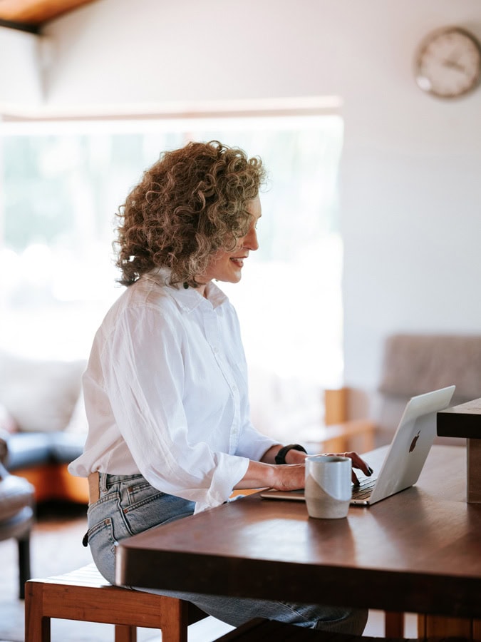 Photo by Florence Charvin Photography of A person with curly hair and a white shirt is sitting at a table using a laptop, with a mug nearby in a bright, indoor setting.