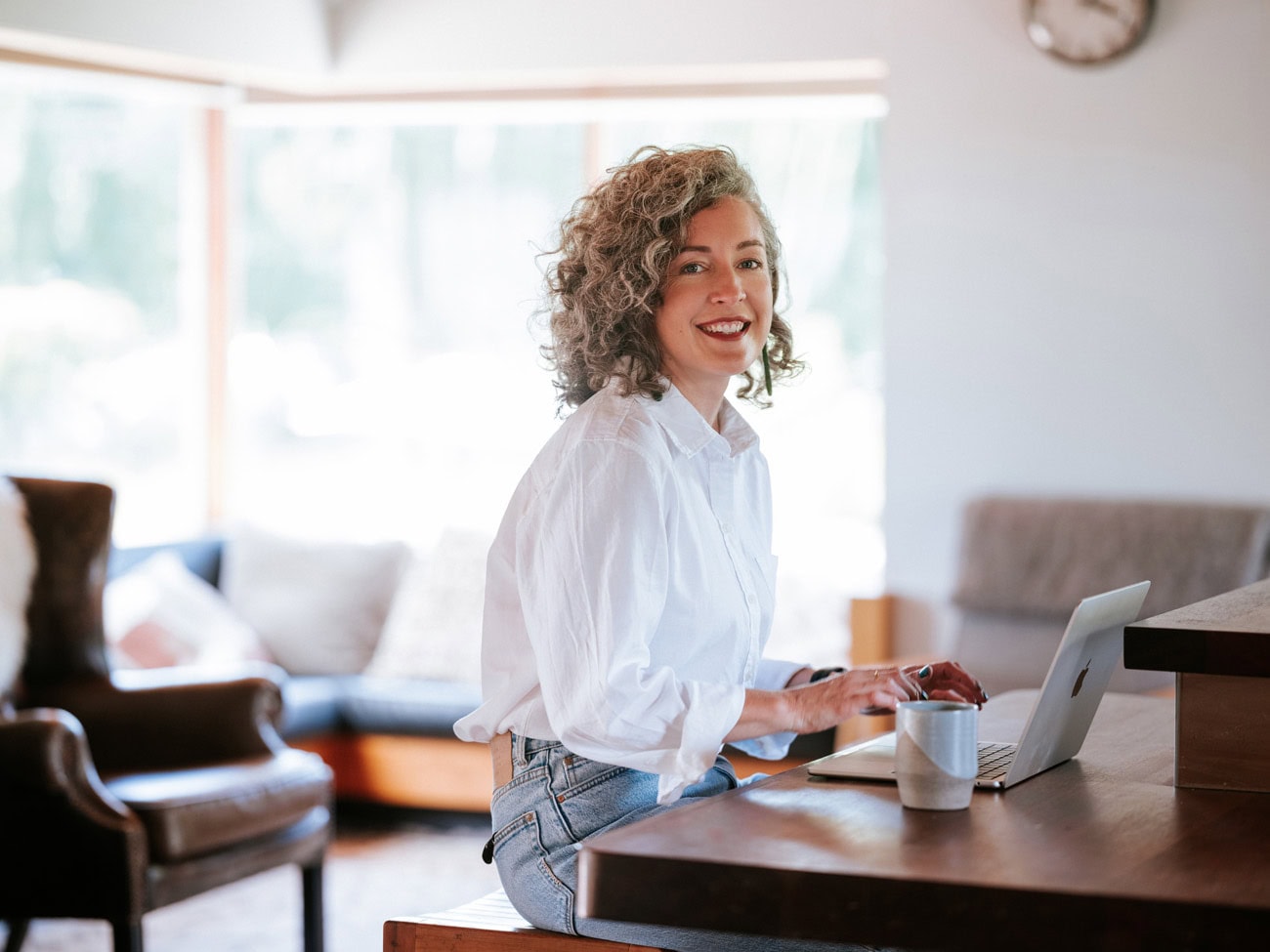 Photo by Florence Charvin Photography of A woman with curly hair wearing a white shirt sits at a table, smiling at the camera whilst using a laptop. A mug is on the table and a sitting room is visible in the background.