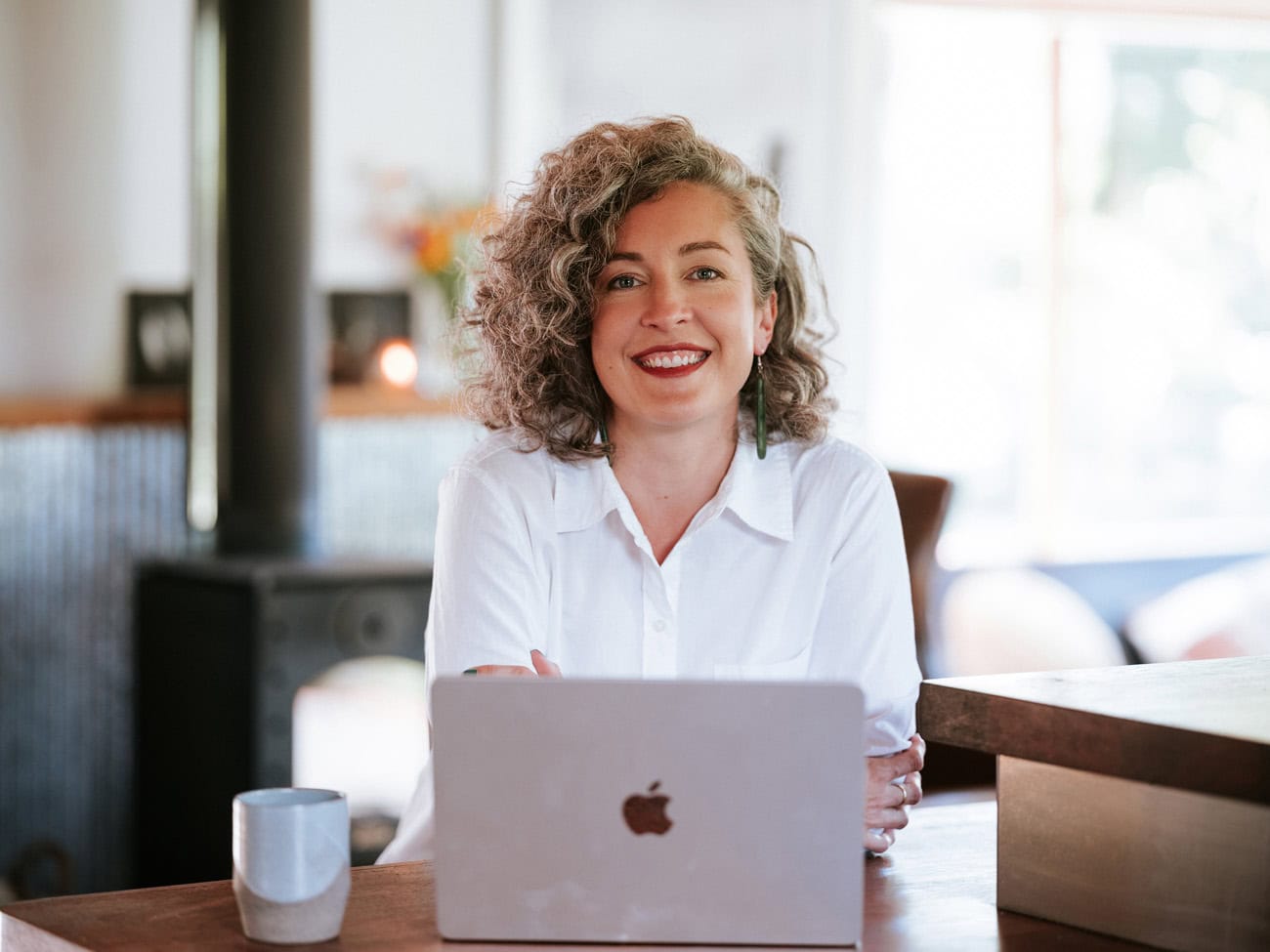 Photo by Florence Charvin Photography of A person with curly hair and a white shirt sits at a table, smiling at the camera, with a laptop and a coffee mug in front of them.